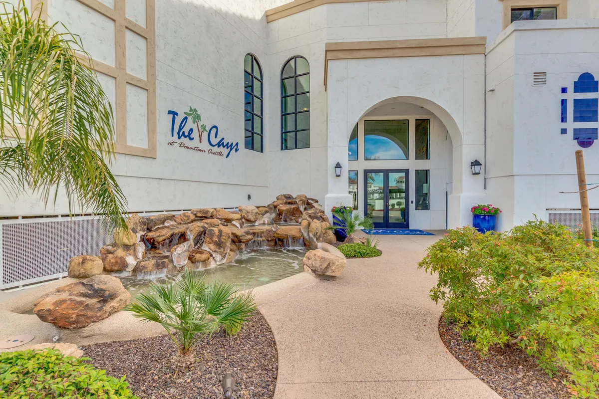 The inviting building entrance, highlighted by a tranquil water feature, lush landscaping, and welcoming signage, creates a sense of arrival and connection to the community.