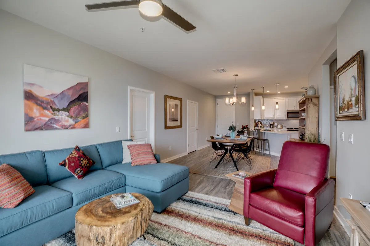 This image captures a comfortable, open-concept living space featuring a blue sectional sofa, a red armchair, a wooden coffee table, wall artwork, and a view of the adjacent dining area. The space looks inviting and ideal for both relaxing and entertaining, embodying the central living area described in the section.