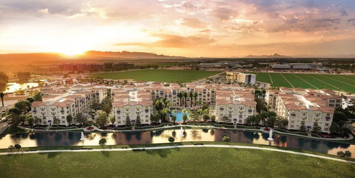 A tranquil exterior view featuring apartment buildings with private balconies overlooking landscaped gardens, a sparkling swimming pool, and water features. The ambiance reflects the beauty of Arizona sunsets and the leisurely lifestyle of the community.
