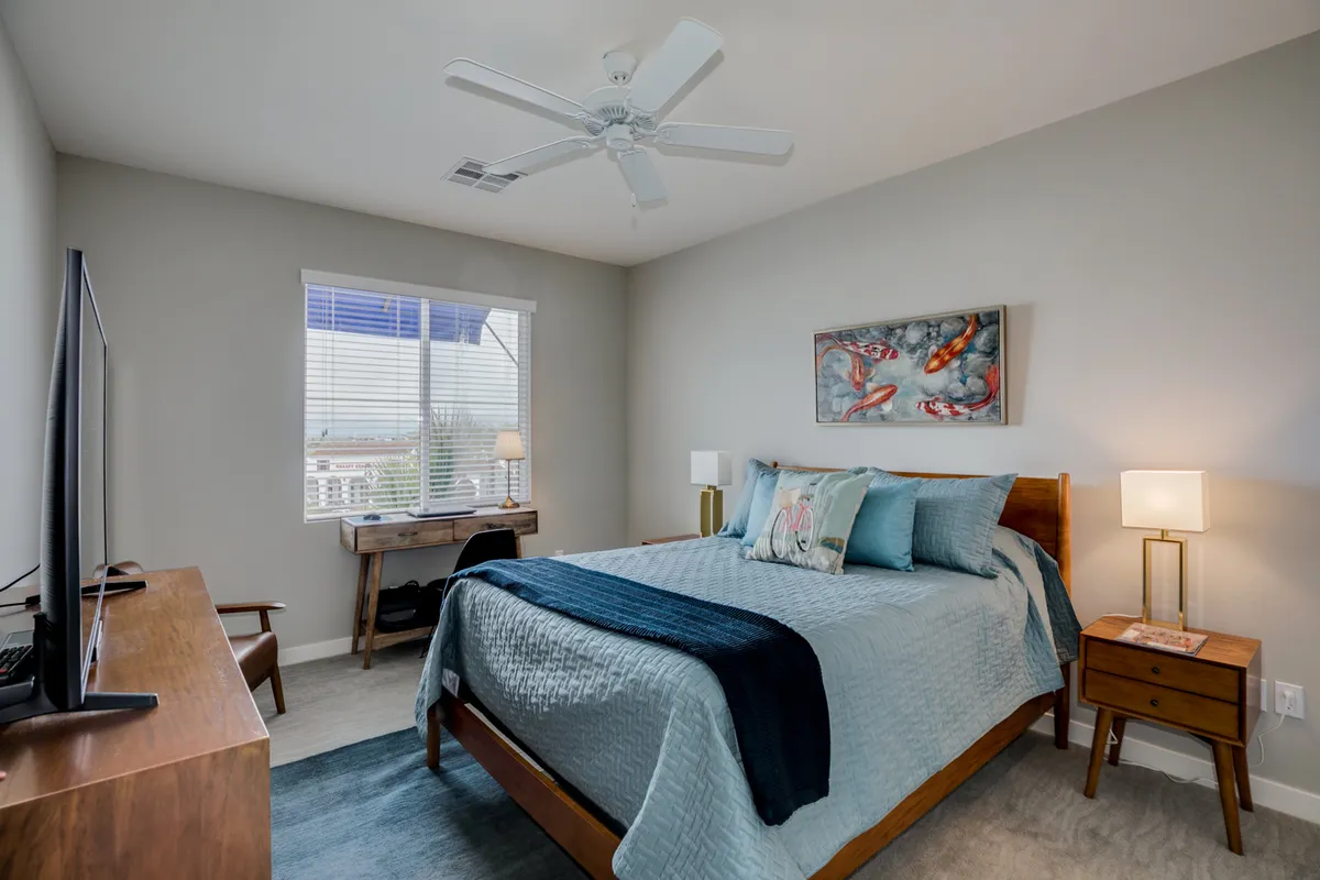 A stylish bedroom featuring a plush bed with crisp linens, a contemporary desk with a lamp for working, a wall-mounted television, and calming artwork. Sunlight streams in, creating a bright and restful atmosphere ideal for both sleep and productivity.