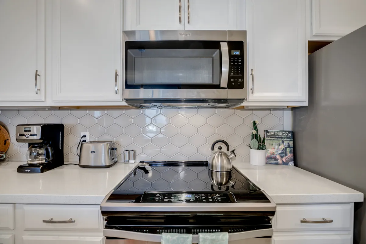 A modern kitchen scene showing stainless steel appliances, a sleek black coffee maker, a contemporary hexagonal tile backsplash, and a decorative kettle. The setting looks ready for morning routines or casual entertaining, capturing a sense of daily comfort and convenience.
