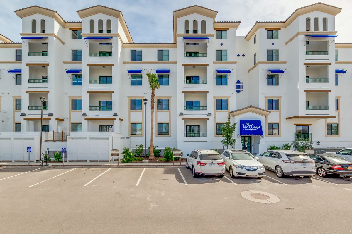 The image displays the exterior facade of The Cays with inviting palm trees, clear signage, and a sunlit atmosphere. The building looks modern and well-maintained, and balconies suggest opportunities for relaxation and fresh air.