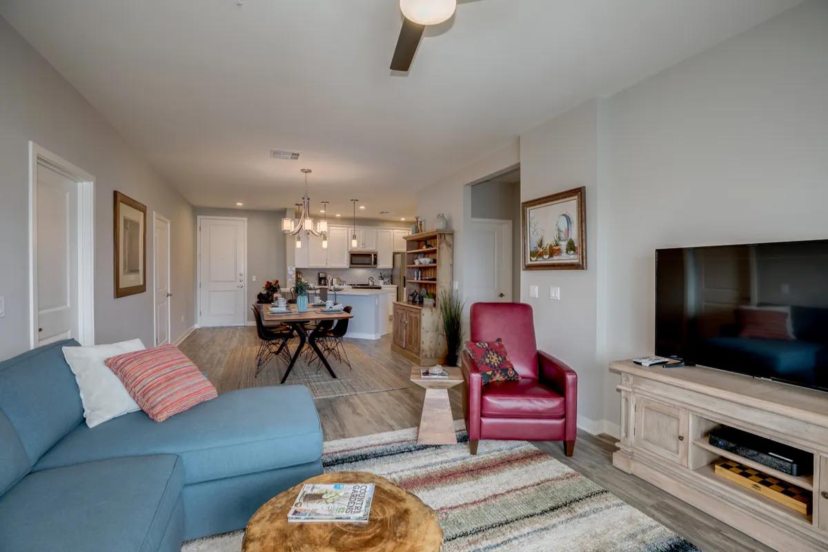 The photo highlights a bright, homey living and dining space. It features a blue sectional sofa, a red accent chair, a wooden coffee table, and a dining area with a chandelier, all bathed in natural light. The layout emphasizes openness and flow between lounging and dining.